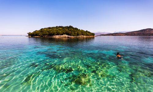 A picture of one of the beautiful islands at Ksamil on the Albanian Riviera. There is a person swimming in the clear blue sea.
